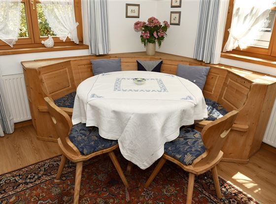 Ein gemütlicher Essbereich mit einer runden Tischgruppe und Holzsitzen. Die Fenster sind mit Vorhängen geschmückt, und auf dem Tisch liegt eine weiße Tischdecke.