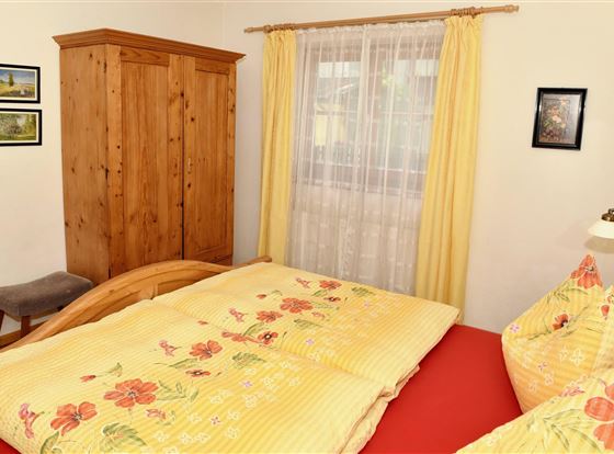 Ein gemütliches Schlafzimmer mit einer Holzkommode und einem großen Bett. Die Bettwäsche ist gelb mit floralen Mustern, und die Fenster sind mit hellen Vorhängen geschmückt.