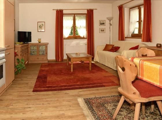 Ein gemütliches Wohnzimmer mit Holzfußboden und einem roten Teppich. Es gibt eine Couch, einen Esstisch und große Fenster mit roten Vorhängen.