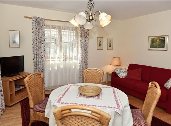 Ein gemütliches Wohnzimmer mit einem roten Sofa und einem Esstisch. Die Fenster sind mit Vorhängen geschmückt und es gibt Kunstwerke an den Wänden.