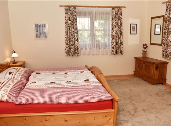 Ein gemütliches Schlafzimmer mit einem Holzbett und karierten Bettdecken. Die Wände sind hell, und Fenster mit floralen Vorhängen sorgen für Tageslicht.