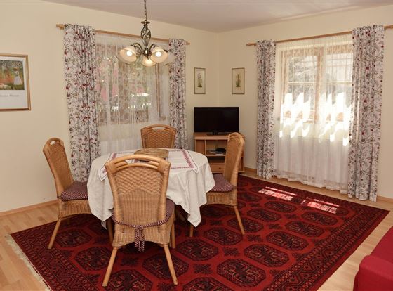 Ein gemütlicher Essbereich mit einem runden Tisch und Stühlen. Der Raum hat helle Wände, Vorhänge mit Blumenmuster und einen Fernseher.