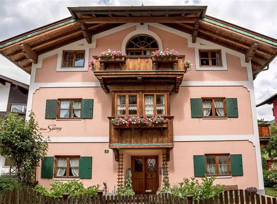 Ein traditionelles bayerisches Haus mit rosa Fassade und Holzbalkonen. Es ist von grünen Pflanzen und Blumen umgeben.