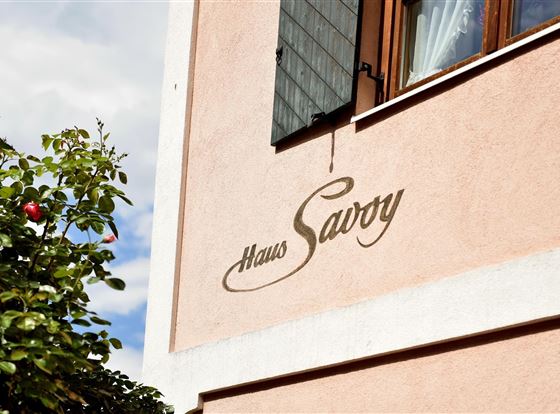 Ein rosa Haus mit einer eleganten Beschriftung "Haus Savvy". Im Vordergrund sind einige grüne Blätter und eine Frucht zu sehen.