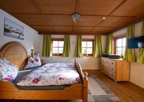 Ein gemütliches Schlafzimmer mit einem großen Holzbett und bunten Kissen. Die Fenster sind mit grünen Vorhängen dekoriert und ein Fernseher hängt an der Wand.