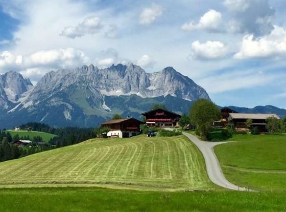 Eine malerische Landschaft mit grünen Wiesen und traditionellen Häusern. Im Hintergrund erheben sich beeindruckende Berge unter einem blauen Himmel.