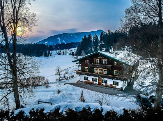 Ein malerisches Alpenhaus im Schnee mit Blick auf die umliegenden Berge. Die Szene wird von einem sanften Sonnenuntergang beleuchtet.