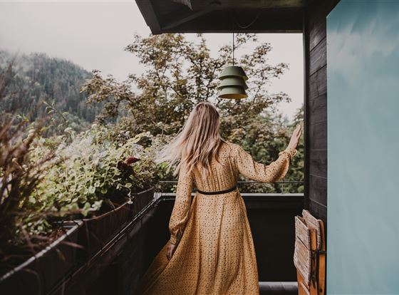 Eine Frau mit langen Haaren steht auf einem Balkon und blickt nach draußen. Im Hintergrund sind grüne Bäume und ein bewölkter Himmel zu sehen.