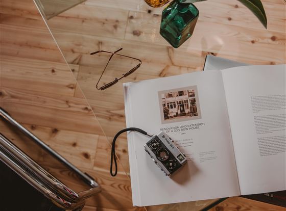 Ein offenes Buch liegt auf einem Glas Tisch, neben einer Kamera und einer Brille. Im Hintergrund steht eine Vase mit Pflanzen.