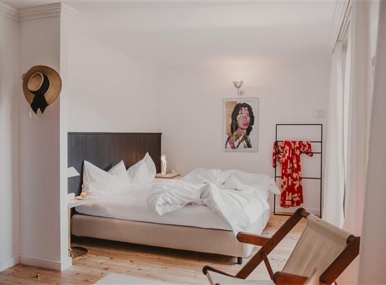 Ein modernes Schlafzimmer mit einem großen Bett und weißen Bettdecken. An der Wand hängt ein Kunstwerk, und ein rotes Kleid hängt im Raum.