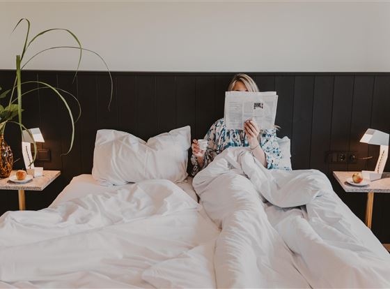 Eine Person sitzt im Bett und liest eine Zeitung. Neben dem Bett stehen kleine Tische mit Lampen und einer Pflanze.