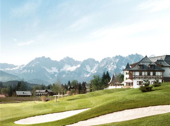 A picturesque landscape with a golf course and an elegant building. Majestic mountains rise in the background under a clear sky.