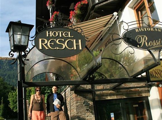 Ein Hotel mit dem Namen "Resch" und einem einladenden Eingang. Zwei Personen gehen die Treppe hinunter, während Blumen die Umgebung schmücken.