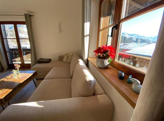 Ein heller Wohnraum mit einem beigen Sofa und einer Holzbank. Auf dem Fensterbrett stehen Blumen und der Blick geht auf Berge und einen blauen Himmel.