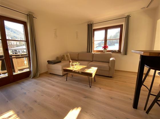 Ein gemütliches Wohnzimmer mit einem hellen Sofa und einem Couchtisch. Große Fenster lassen viel Licht herein und bieten einen Blick nach draußen.