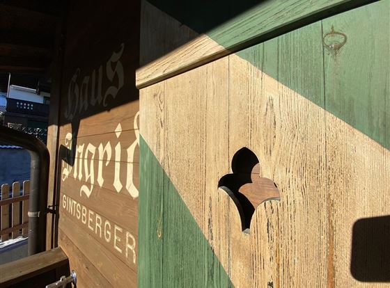 Ein Holzhaus mit einer rustikalen Fassade und einem hölzernen Detail in Form eines stilisierten Fensters. Die Farben sind überwiegend braun und grün, mit sanften Licht- und Schattenspielen.