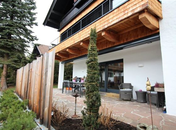 Eine moderne Terrasse mit einem Garten und einem Holzsichtschutz. Im Hintergrund ist ein gemütlicher Tisch und Stühle zu sehen.