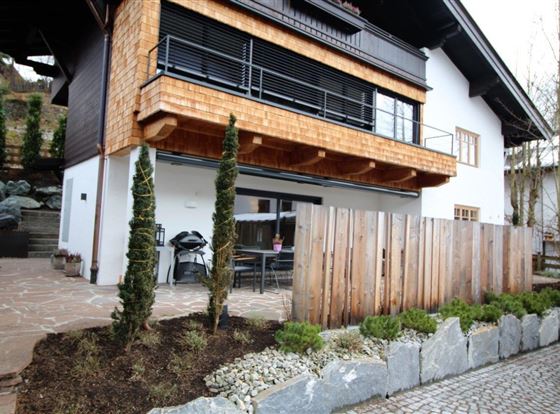 Ein modernes Haus mit einem Holzbalkon und einem gepflegten Vorgarten. Der Außenbereich ist mit Steinen und Pflanzen gestaltet, und es gibt eine Terrasse mit Sitzmöglichkeiten.
