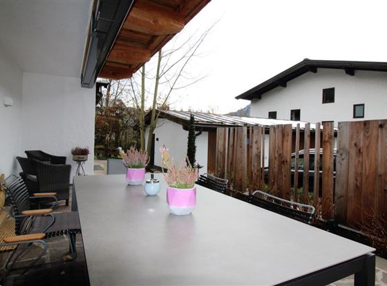 Eine Terrasse mit einem langen Tisch und bunten Töpfen mit Pflanzen. Im Hintergrund sind moderne Häuser und ein Zaun aus Holz zu sehen.