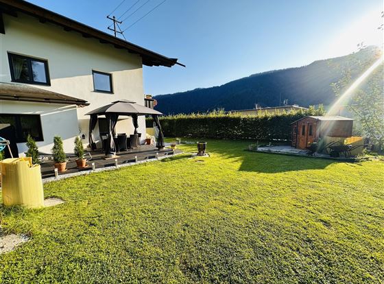 Ein gepflegter Garten mit grünem Rasen und einer Pergola. Im Hintergrund sind Berge und ein schöner blauer Himmel zu sehen.