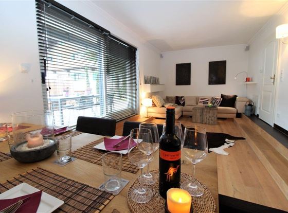 Ein modernes Wohnzimmer mit Holzfußboden und großen Fenstern. Der Esstisch ist festlich gedeckt mit Weinglas und einer Flasche Wein.
