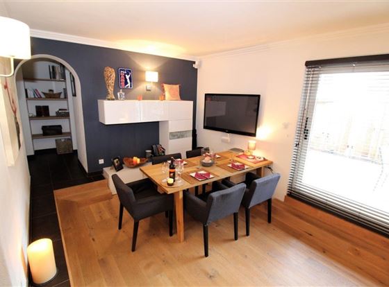 Ein modernes Esszimmer mit einem Holztisch und schwarzen Stühlen. Im Hintergrund befindet sich ein Fernseher und ein gemütlicher Wohnbereich mit Zugang zu einem Fenster.