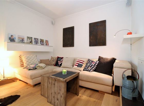 Ein gemütliches Wohnzimmer mit einem großen beigen Sofa und bunten Kissen. Neben dem Sofa steht ein Holztisch und an der Wand hängen einige Bilder.