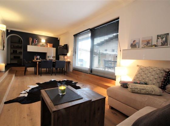 Ein modernes Wohnzimmer mit einem gemütlichen Sofa, einem Holzkaffee-Tisch und einem Esstisch im Hintergrund. Große Fenster sorgen für viel Tageslicht und eine einladende Atmosphäre.
