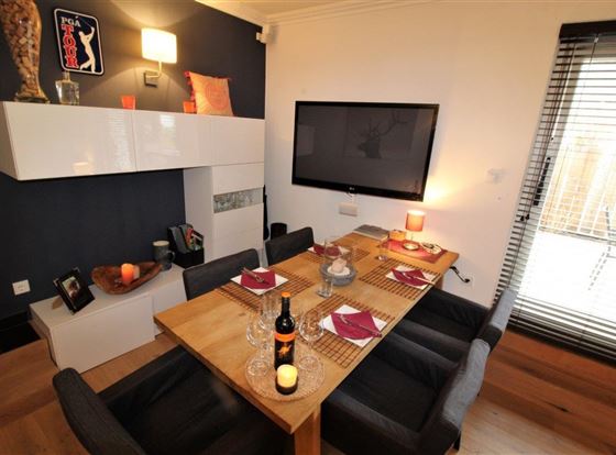 Ein modernes Esszimmer mit einem großen Holztisch und stilvollen Stühlen. Die Wände sind dunkel, und ein Fernseher hängt über dem Tisch.