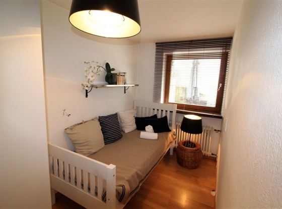 Ein gemütliches Zimmer mit einem Bett, einem Fenster und einer warmen Lampe. Die Wände sind hell und es gibt eine kleine Pflanze auf einem Regal.