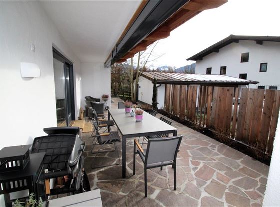 Eine einladende Terrasse mit einem großen Tisch und Stühlen. Hinter der Terrasse ist ein Garten mit einem Holzzaun und einem daneben stehenden Gebäude.