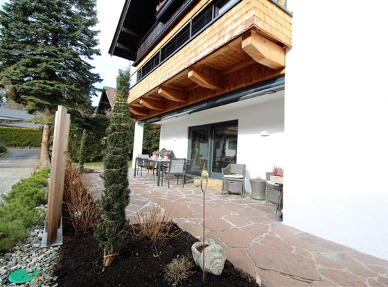 Eine moderne Terrasse mit einem Essbereich und einem klaren Blick auf eine Gartenlandschaft. Der Bereich ist von grünen Pflanzen und Steinen umgeben.
