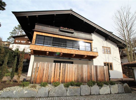 Ein modernes Haus mit einem Holzüberhang und großen Fenstern. Der Eingangsbereich ist von einer Holzlattenzaun und Steinen umgeben.
