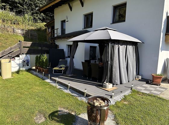 Eine gemütliche Terrasse mit einem grauen Pavillon und Gartenmöbeln. Im Hintergrund steht ein Haus und der Rasen ist gut gepflegt.