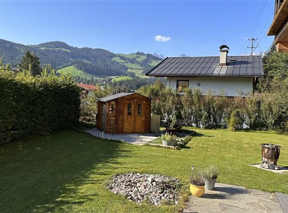 Ein schöner Garten mit einem Holzschuppen und gepflegtem Rasen. Im Hintergrund sind grüne Berge und ein blauer Himmel zu sehen.