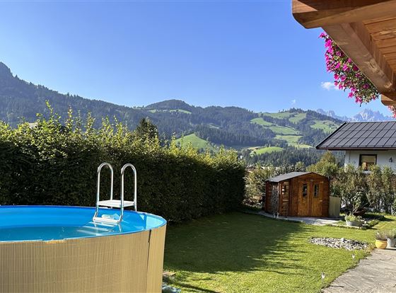 Ein schöner Garten mit einem Aufstellpool und einer malerischen Berglandschaft im Hintergrund. Der Himmel ist klar und die Sonne scheint.