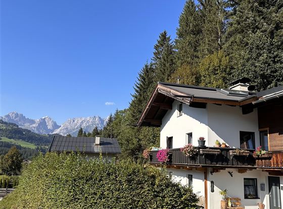 Ein schönes Haus mit Balkon in einer grünen Umgebung. Im Hintergrund sind majestätische Berge und ein klarer blauer Himmel zu sehen.