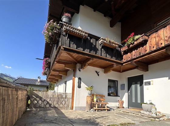 Ein charmantes Haus mit einem Holzbalkon und bunten Blumen. Im Vordergrund gibt es einen kleinen Sitzbereich und viel Tageslicht.