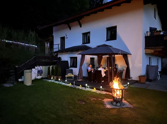 Ein gemütliches Zuhause mit einem großen Garten und einem offenen Feuer. Im Hintergrund sitzen Menschen unter einem Sonnenschirm und genießen die Abendstimmung.