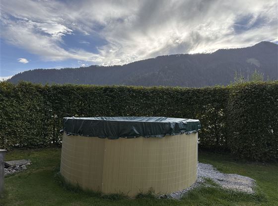 Ein runder Pool mit grüner Abdeckung steht auf einer Wiese. Im Hintergrund sind Hügel und ein wolkenverhangener Himmel zu sehen.
