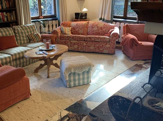 Ein gemütliches Wohnzimmer mit mehreren Sofas in warmen Farben. Der Raum hat große Fenster und einen Kamin, der eine einladende Atmosphäre schafft.