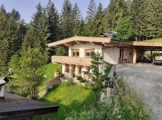 Ein gemütliches Haus in den Bergen mit einer Veranda und einer Garage. Umgeben von grünen Wiesen und hohen Bäumen.