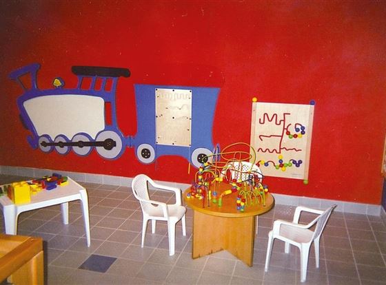 Ein bunter Spielbereich für Kinder mit einem Zug-Motiv an der Wand. Es gibt einen runden Tisch mit Spielzeug und Stühlen.