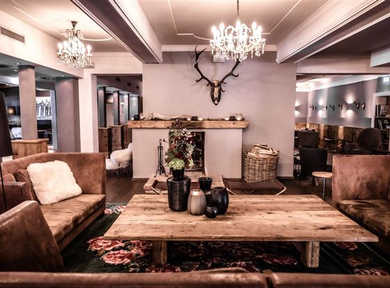 Eine stilvolle Lounge mit gemütlichen Sofas und einem Holztisch. An der Wand hängt ein Geweih, und mehrere Kronleuchter sorgen für eine angenehme Beleuchtung.