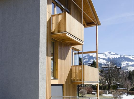 Ein modernes Gebäude mit Holzverkleidung und großen Fenstern. Im Hintergrund sind schneebedeckte Berge und ein klarer Himmel zu sehen.