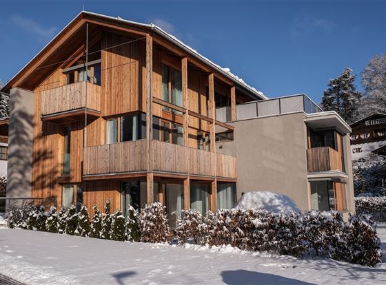 Ein modernes Haus mit Holzfassade und großen Fenstern. Rundherum liegt eine Schneedecke, und der Himmel ist klar und blau.