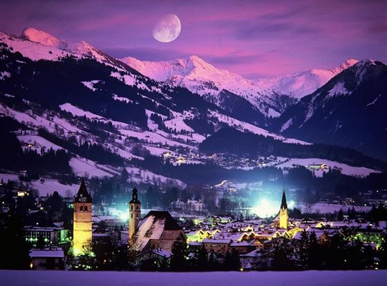 Eine malerische Berglandschaft bei Nacht, mit verschneiten Gipfeln und einem klaren Mond. Im Vordergrund ist eine charmante Stadt mit historischen Gebäuden und sanften Lichtern zu sehen.