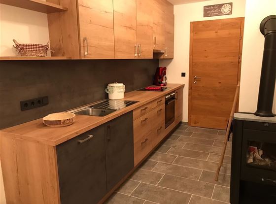 Eine moderne Küche aus Holz mit grauen Schränken und einem hellen Boden. Die Ausstattung umfasst eine Spüle, einen Herd und einen Holzofen.