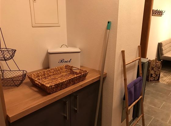 Eine gemütliche Ecke mit einem Holzregal und einem Brotkasten. Neben dem Regal befindet sich ein Wäschegestell und ein Korb auf der Arbeitsfläche.