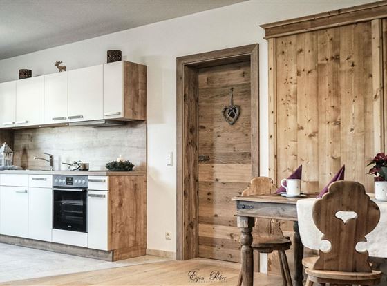 Eine moderne Küche mit hellen, schlichten Möbeln und Holzelementen. An einem kleinen Tisch stehen zwei Stühle und eine blühende Pflanze.